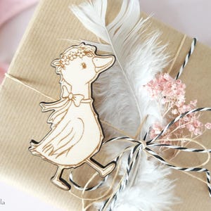 May include: A wooden duck ornament with a flower crown, a bow, and a white feather. The duck is on a brown paper gift bag with white and black twine and pink flowers.