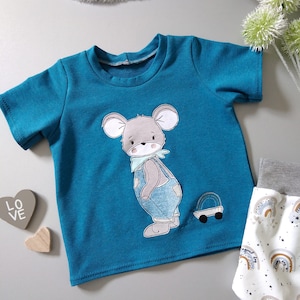 May include: A teal blue short-sleeved t-shirt with an embroidered illustration of a grey mouse wearing overalls and pulling a toy wagon. The mouse is looking to the right.