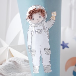 May include: A light blue cone-shaped item with an embroidered astronaut design. The astronaut is wearing a white spacesuit with silver accents, a helmet, and waving. The astronaut has brown hair and a smiling face. The design is on a light blue background.