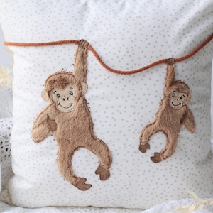 Peut inclure: Un coussin blanc avec un motif de petits points gris. Deux appliqués de singes bruns sont suspendus à une corde marron. Les singes ont des visages souriants et sont faits d'une matière douce et texturée.