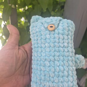 May include: A light blue crochet pouch made of soft, textured yarn. The rectangular pouch features a small, round, light-coloured wooden button closure at the top centre and a small, round, matching blue crochet pom-pom attached to the side.