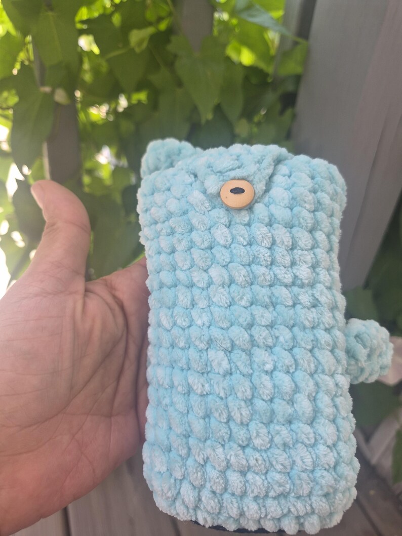 May include: A light blue crochet pouch made of soft, textured yarn. The rectangular pouch features a small, round, light-coloured wooden button closure at the top centre and a small, round, matching blue crochet pom-pom attached to the side.