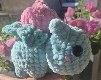 May include: A handmade plush toy resembling a cartoon character. It's mainly light blue with a pink top, a black button eye, and a small pink nose. The toy is crafted from a soft, textured material, giving it a cuddly appearance.