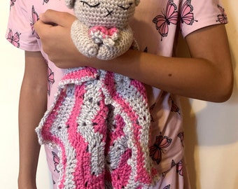 May include: A grey crocheted stuffed animal with closed eyes, holding a pink crocheted blanket. The stuffed animal is being held by a person wearing a pink shirt with a butterfly pattern.