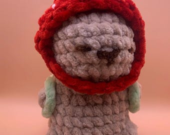May include: A handmade crochet doll with a red hood and a light green accent. The doll is grey with brown facial features. The doll is a cute, whimsical design.