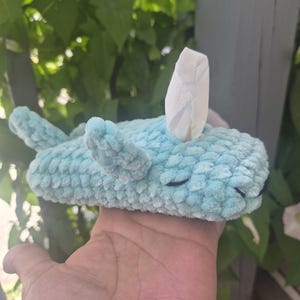 May include: A light blue crocheted tissue box cover shaped like a whale. The plush, textured yarn creates a soft, rounded body with small fins on the sides and a tail at the back. A single white tissue protrudes from the top of the whale's back.