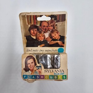 May include: Vintage Sylvania Blue Dot Flashcubes package. The package features a family photo and a woman holding a camera. Text on the package reads "don't miss any memories" and "FLASHCUBES". The package is beige with blue and green accents.