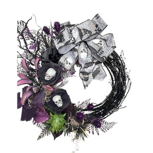 May include: A Halloween wreath with a black twig base, adorned with black roses, purple foliage, and a gray bow featuring skull patterns. The wreath includes small white skull accents and various decorative elements.