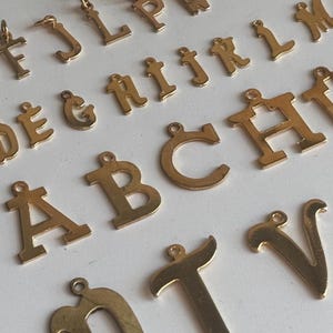 May include: A collection of gold-colored metal alphabet charms, both uppercase and lowercase, scattered on a white surface. Each letter has a small loop at the top for attaching to a chain or bracelet. The letters are in a classic serif font.
