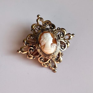 May include: A vintage cameo brooch with a detailed profile carving of a woman. The cameo is set in a gold-toned filigree frame with an ornate, floral design. The brooch is a classic piece of jewelry.