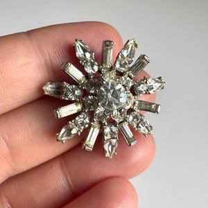 May include: A vintage silver-toned brooch in a starburst design. The brooch features a large, round, clear crystal center surrounded by smaller, clear, rectangular and pointed crystals. The crystals are set in a silver metal frame.
