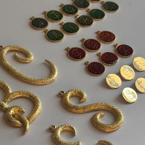 May include: An assortment of gold-colored, textured metal pendants and charms. The charms include individual letters in green and red enamel, and gold-colored circular charms with engraved letters. Several large, textured gold-colored letter pendants are also present.