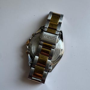 May include: A two-tone gold and silver Citizen watch with a chronograph function. The watch has a stainless steel strap with gold accents. The back of the watch is visible, showing the Citizen logo and the words "Water Resist 5 Bar" and "Japan Movt."