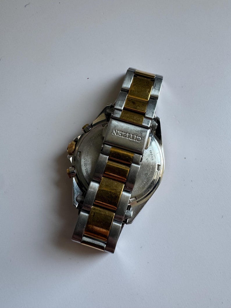 May include: A two-tone gold and silver Citizen watch with a chronograph function. The watch has a stainless steel strap with gold accents. The back of the watch is visible, showing the Citizen logo and the words "Water Resist 5 Bar" and "Japan Movt."