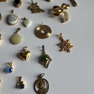 May include: A collection of gold-tone pendants with various designs, including a heart, a seashell, a snowflake, a cross, a crown, a record, and a figure of a person. Some pendants feature gemstones, such as blue, green, and purple.