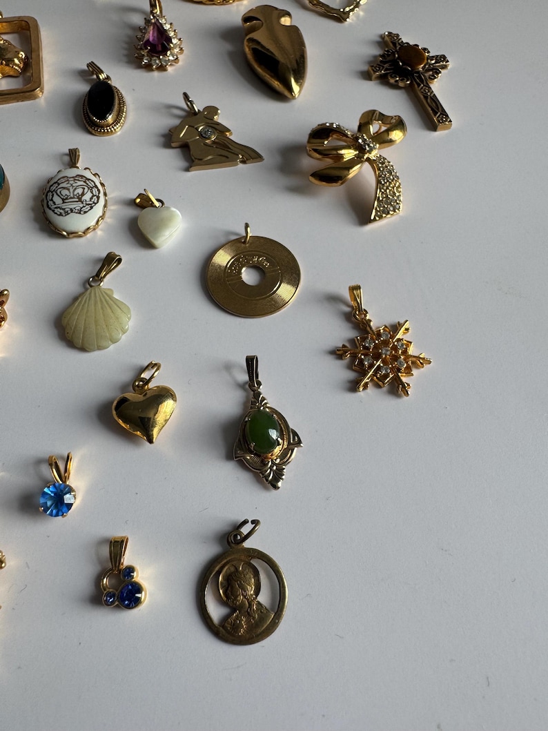 May include: A collection of gold-tone pendants with various designs, including a heart, a seashell, a snowflake, a cross, a crown, a record, and a figure of a person. Some pendants feature gemstones, such as blue, green, and purple.
