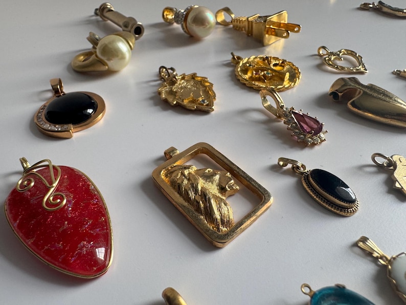 May include: A collection of gold and silver pendants with various designs, including a lion, a heart, a crown, and a stone. The pendants are on a white surface.