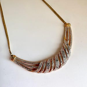 May include: A gold-toned necklace with a curved, textured pendant. The pendant features a swirling design with rows of clear and black gemstones. The necklace chain is a delicate gold color, and the overall design is elegant and eye-catching.