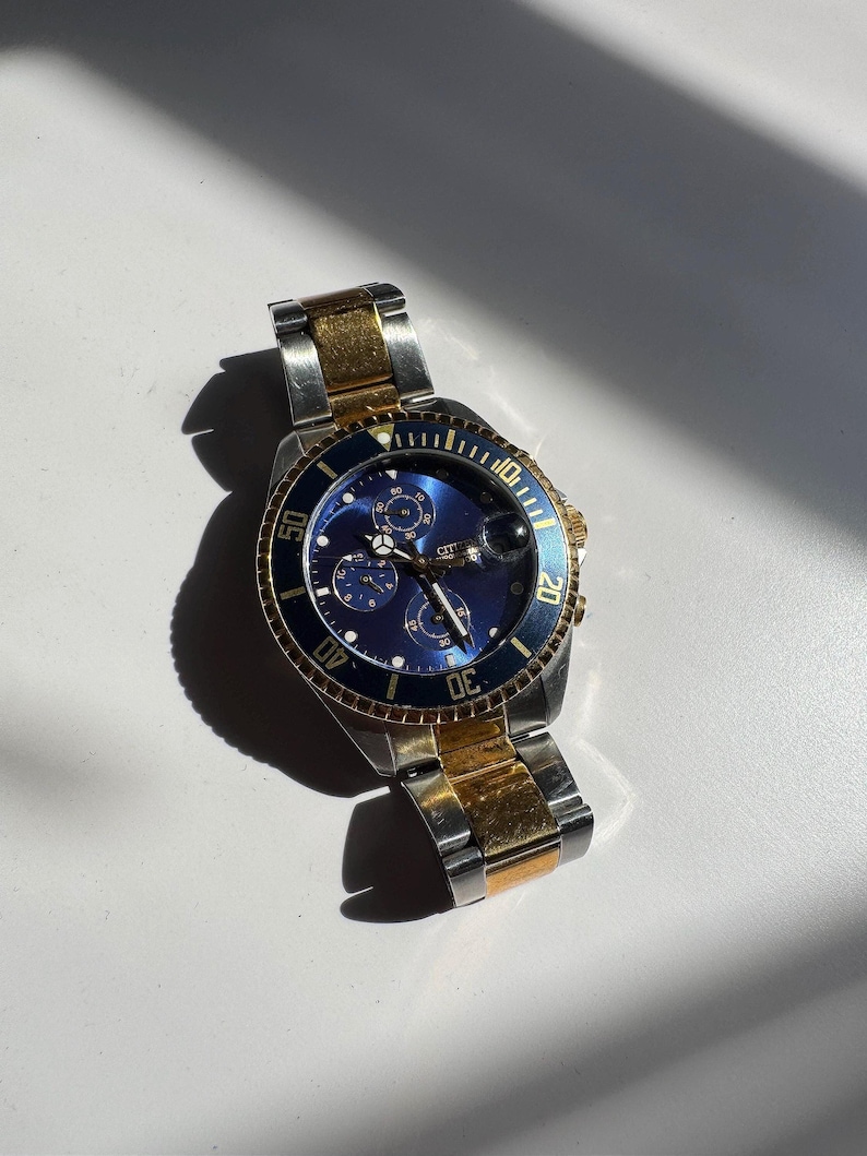 May include: A two-tone gold and silver chronograph wristwatch with a blue face and a rotating bezel. The watch has a date window at the 4 o'clock position and a Citizen logo.