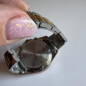 May include: A two-tone silver and gold wristwatch with a chronograph face and a stainless steel strap. The watch has a silver face with black markings and hands. The strap is made of alternating silver and gold links.