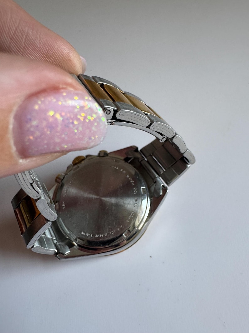 May include: A two-tone silver and gold wristwatch with a chronograph face and a stainless steel strap. The watch has a silver face with black markings and hands. The strap is made of alternating silver and gold links.