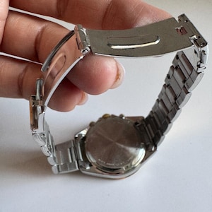 May include: A silver-toned wristwatch with a stainless steel strap and a round face. The watch is being held by a hand.