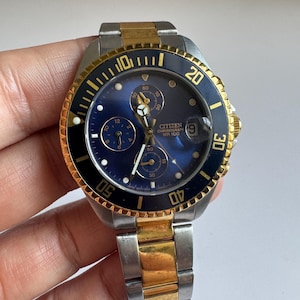 May include: A two-tone gold and silver Citizen chronograph wristwatch with a blue face and black bezel. The watch has a date window at the 3 o'clock position and a chronograph function. The watch is water resistant to 100 metres.