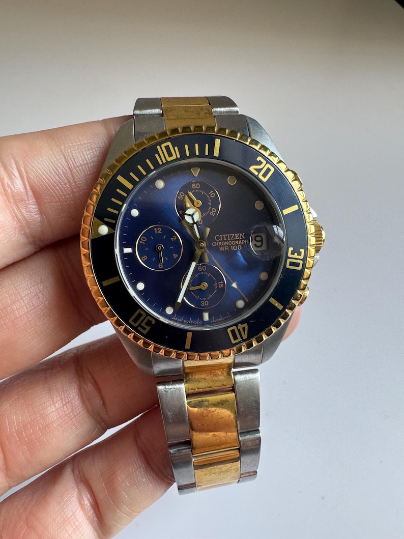 May include: A two-tone gold and silver Citizen chronograph wristwatch with a blue face and black bezel. The watch has a date window at the 3 o'clock position and a chronograph function. The watch is water resistant to 100 metres.
