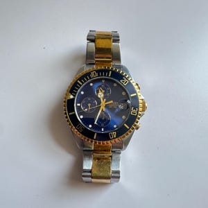 May include: A two-tone gold and silver wristwatch with a blue face and a black bezel. The watch has a chronograph function and a date window.