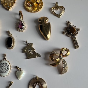 May include: A collection of gold-tone charms with various designs, including a heart, a cross, a bow, a record, a seashell, a crown, a tree, a teardrop, a mermaid, and an arrow. The charms are arranged on a white surface.