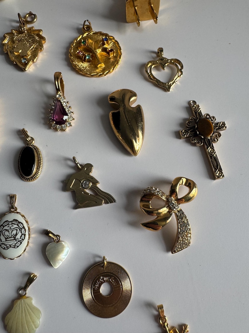 May include: A collection of gold-tone charms with various designs, including a heart, a cross, a bow, a record, a seashell, a crown, a tree, a teardrop, a mermaid, and an arrow. The charms are arranged on a white surface.