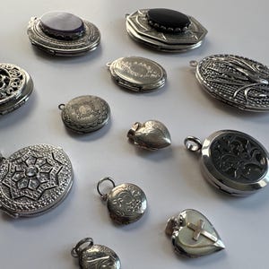 Op de afbeelding: Een verzameling van 11 zilveren hanger-medaillons, sommige met ingewikkelde ontwerpen, andere met eenvoudige gravures. De medaillons zijn in verschillende vormen en maten, waaronder rond, ovaal, hartvormig en achthoekig.