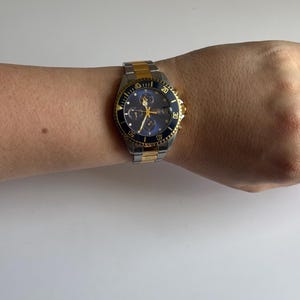 May include: A two-tone gold and silver wristwatch with a black face and a blue bezel. The watch has a chronograph function and a date window.
