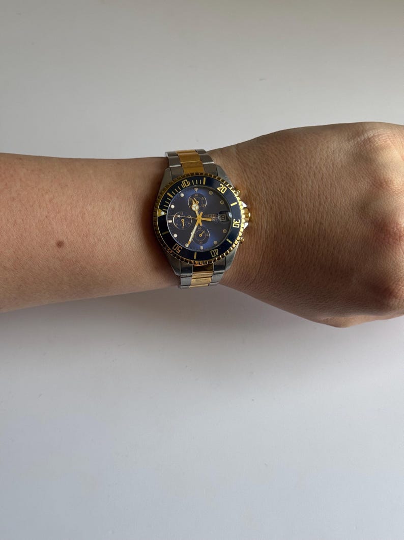 May include: A two-tone gold and silver wristwatch with a black face and a blue bezel. The watch has a chronograph function and a date window.