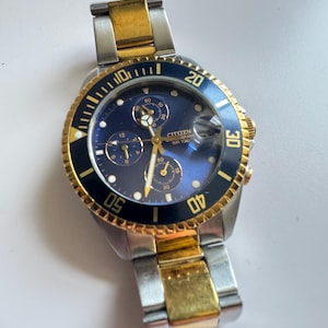 May include: A two-tone gold and silver Citizen chronograph wristwatch with a blue face and black bezel. The watch has a date window and a rotating bezel with markings for minutes. The text "CITIZEN GRAPH WR 100" is printed on the face.