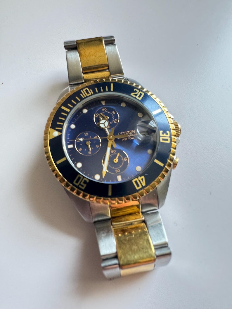 May include: A two-tone gold and silver Citizen chronograph wristwatch with a blue face and black bezel. The watch has a date window and a rotating bezel with markings for minutes. The text "CITIZEN GRAPH WR 100" is printed on the face.