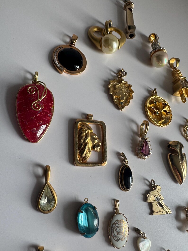 May include: A collection of gold-tone pendants with various gemstones and designs, including a heart-shaped pendant with a red stone, a lion pendant, a pendant with a blue stone, and a pendant with a pearl.