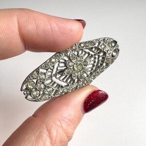 May include: An oval-shaped, silver-toned brooch with a floral and geometric design. The brooch is encrusted with small, clear rhinestones, and is held by a hand with red nail polish.