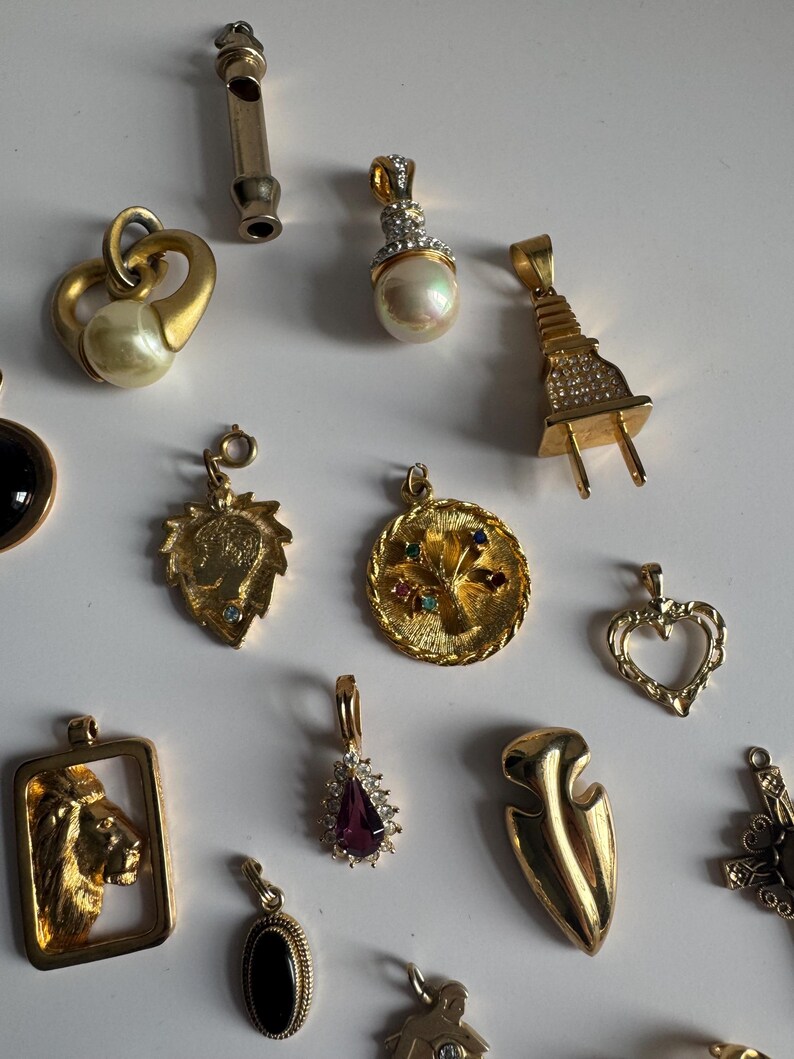 May include: A collection of 15 gold-tone charms, including a lion, a heart, a tree, a plug, and a whistle. The charms are all different sizes and shapes and have various embellishments, such as rhinestones, pearls, and enamel.
