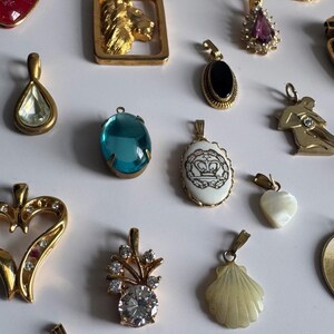 May include: A collection of gold-tone pendants with various gemstones and designs, including a heart, a shell, a flower, a lion, and a crown. The pendants are arranged on a white surface.