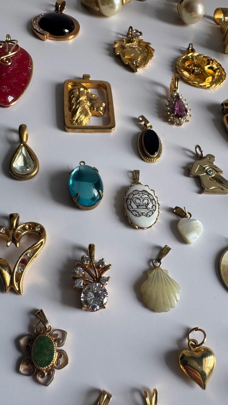 May include: A collection of gold-tone pendants with various gemstones and designs, including a heart, a shell, a flower, a lion, and a crown. The pendants are arranged on a white surface.