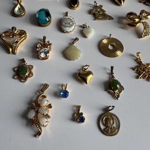 May include: A collection of gold-tone pendants with various designs, including a heart, a seashell, a crown, a flower, a cross, and a blue stone. The pendants are arranged on a white surface.