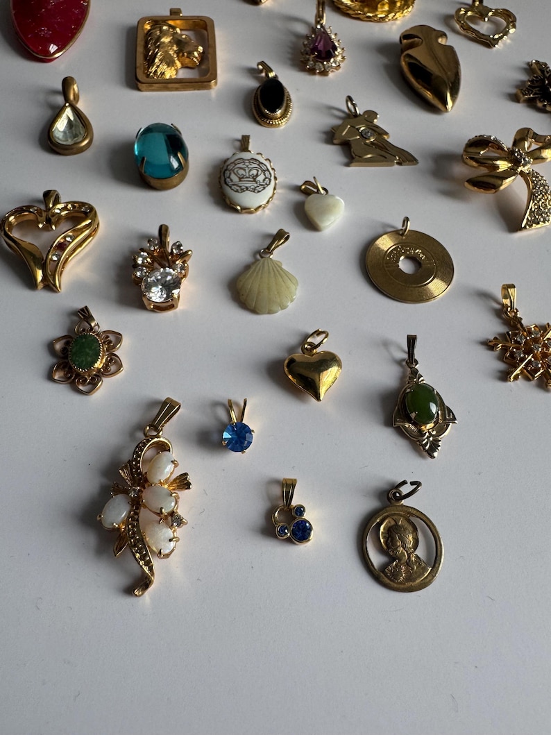 May include: A collection of gold-tone pendants with various designs, including a heart, a seashell, a crown, a flower, a cross, and a blue stone. The pendants are arranged on a white surface.