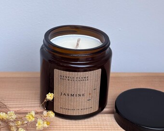 Może przedstawiać: Brązowy szklany słoik z białą świecą sojową w środku. Etykieta zawiera napis "Gentle Flame Soy Wax Candle" i "Jasmine" z małą brązową kropką. Etykieta zawiera również napis "Handmade in Belgium".