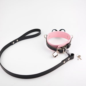 May include: A pink and black collar featuring two small black cat ears on top, a silver-toned metal padlock, and a matching black leash. The set includes two small silver keys. The collar has a buckle closure and silver-toned hardware throughout.