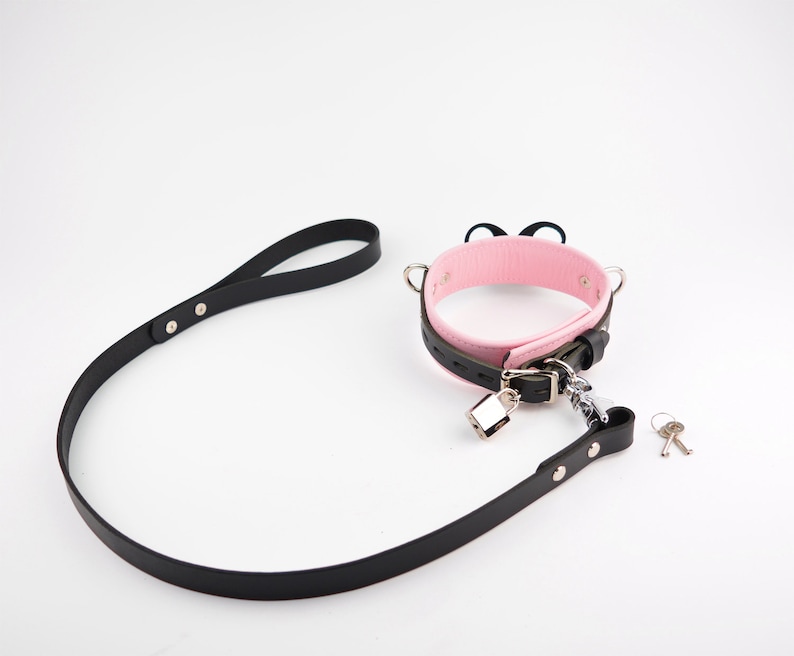 May include: A pink and black collar featuring two small black cat ears on top, a silver-toned metal padlock, and a matching black leash. The set includes two small silver keys. The collar has a buckle closure and silver-toned hardware throughout.