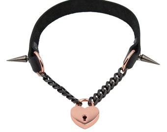 May include: A black leather choker with a rose gold heart-shaped padlock and black metal chain. The choker has two spikes on either side.
