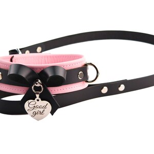 May include: A pink and black collar featuring a black leather strap, a pink inner lining, silver-toned metal hardware, a black bow, and a heart-shaped silver charm engraved with the text Good girl, attached to a long black leather leash.