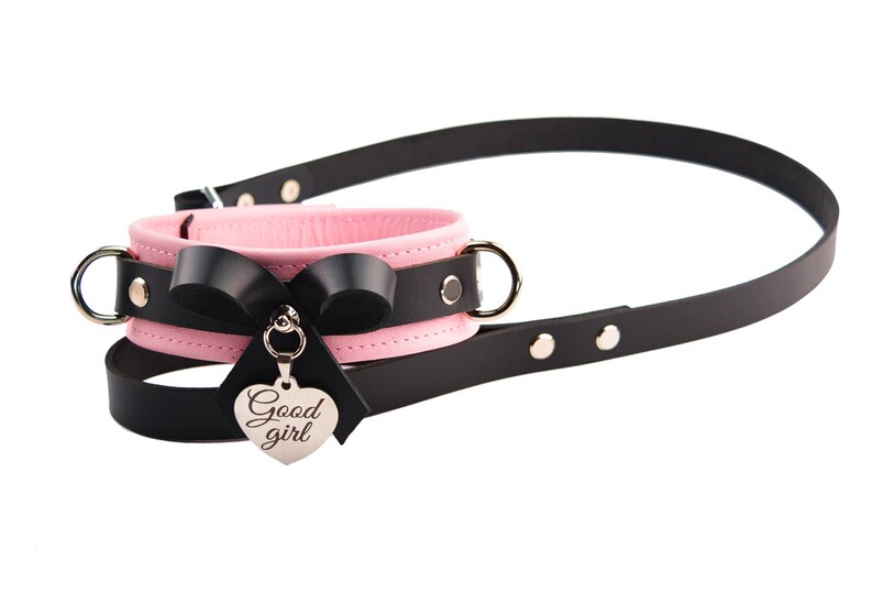 May include: A pink and black collar featuring a black leather strap, a pink inner lining, silver-toned metal hardware, a black bow, and a heart-shaped silver charm engraved with the text Good girl, attached to a long black leather leash.