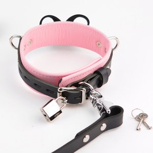 May include: A black and pink collar featuring a silver-toned metal padlock, a matching leash attachment, and two small keys. The collar has a pink interior lining, a black exterior strap with decorative cutouts, and two silver metal D-rings on the sides.